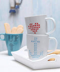 Front view of a set of two white ceramic mugs with blue and red cross-stitch patterns, stacked on a white tray.