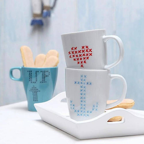 Front view of a set of two white ceramic mugs with blue and red cross-stitch patterns, stacked on a white tray.