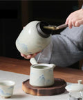 Top Shelf Ceramic Tea Canister With Airtight Lock Lid pouring loose tea into a teapot on a wooden table.