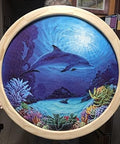 Hand holding a circular wooden frame with a dolphin and coral reef painting, illustrating potential use for No Fuss Ellipse Router Circle Cutting Jig For Woodworking, Easy To Use Circle And Ellipse Routing Jig With Guide Arm, Plunge Router Compatibility, Jigsaw Guide, Dovetail Keys, And More.