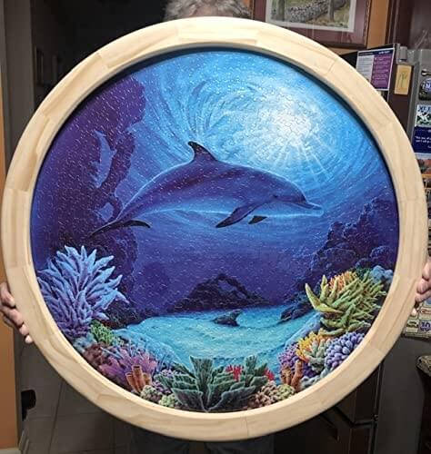 Hand holding a circular wooden frame with a dolphin and coral reef painting, illustrating potential use for No Fuss Ellipse Router Circle Cutting Jig For Woodworking, Easy To Use Circle And Ellipse Routing Jig With Guide Arm, Plunge Router Compatibility, Jigsaw Guide, Dovetail Keys, And More.