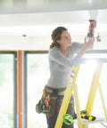 Front view of a woman on a yellow ladder using a cordless drill to cut a hole in a drywall ceiling.