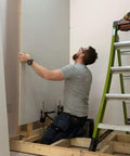 Viking Arm handheld jack/clamp lifting drywall sheet; worker in gray shirt beside a ladder in a framed room.