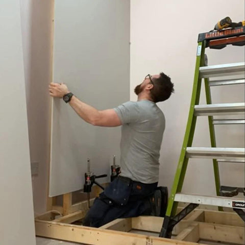 Viking Arm handheld jack/clamp lifting drywall sheet; worker in gray shirt beside a ladder in a framed room.