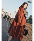 Side view of a woman wearing a long brown coat and black boots, walking in an outdoor setting with palm trees.