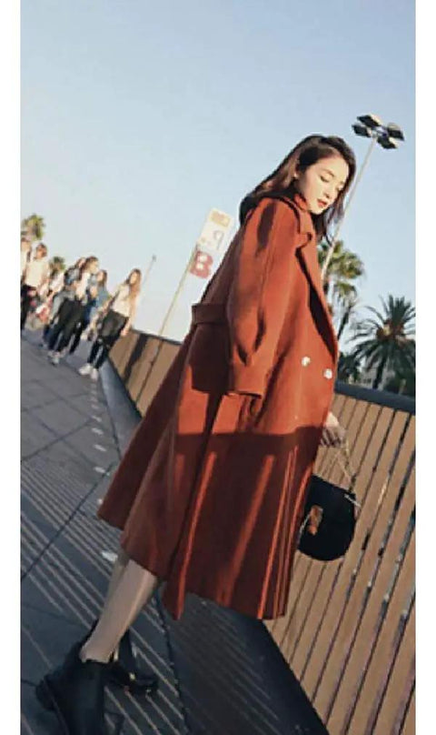 Side view of a woman wearing a long brown coat and black boots, walking in an outdoor setting with palm trees.