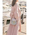 Woman wearing a pink coat and carrying a gray bag, walking by boats at a marina.