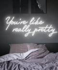 Bedroom wall with 'You're like really pretty' neon light above unmade bed, highlighting a cozy and modern decor ambiance.