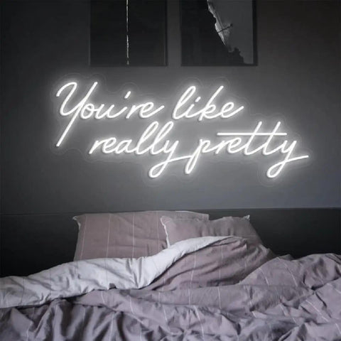 Bedroom wall with 'You're like really pretty' neon light above unmade bed, highlighting a cozy and modern decor ambiance.