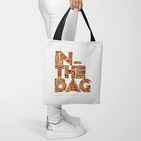 Person holding a tote bag with colorful "IN THE BAG" text design, wearing white pants and sneakers.