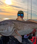 Angler on a boat at sunset holding a large grey offshore saltwater fish; wearing a cap and blue mirrored sunglasses; fishing rods in background