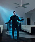 Man standing under ceiling fan indoors, illuminated by light beam, next to modern furniture in dark room with art on wall.
