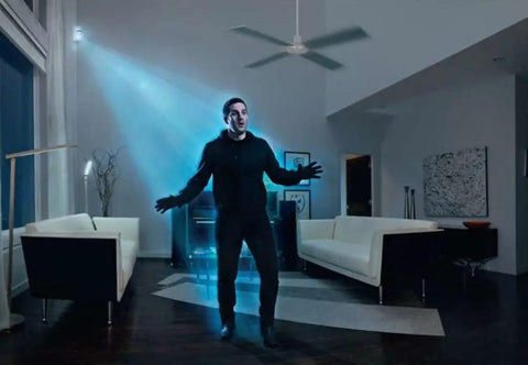 Man standing under ceiling fan indoors, illuminated by light beam, next to modern furniture in dark room with art on wall.