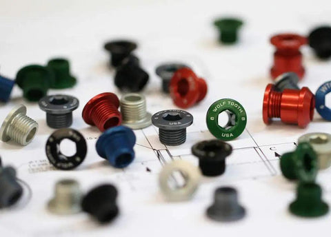 Assorted precision machined chainring bolts and nuts for 1x drivetrains, including 4 mm hex bolt and 6 mm hex nut, on a blueprint.