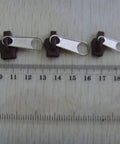 Silver zipper sliders from Six Piece Black Universal Instant Zipper Repair Kit with ruler measuring size on wooden surface.
