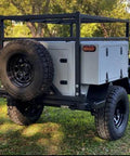 Off-road utility trailer shown from rear angle with black steel frame, spare tire, and gray storage boxes.