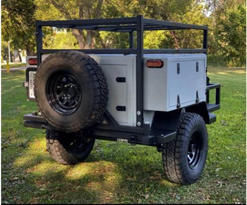 Off-road utility trailer shown from rear angle with black steel frame, spare tire, and gray storage boxes.