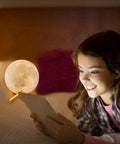Girl in a cozy bedroom reading near a Lit Moon Lamp 3D Printed LED Night Light With 16 Color RGB Modes Remote And Touch Control Adjustable Brightness USB Recharge Realistic 6 Inch Moon Decor For Kids And Birthday Gift.