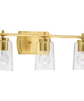 Adley Collection 3 Light Clear Glass Bath Vanity Light in Satin Brass with crystal clear glass shades installed above bathroom mirror