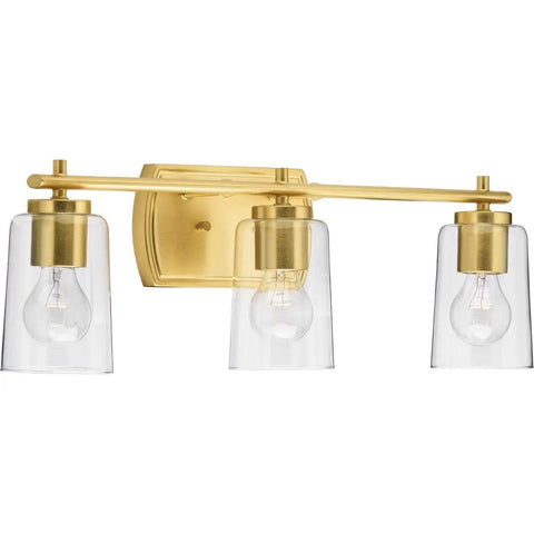 Adley Collection 3 Light Clear Glass Bath Vanity Light in Satin Brass with crystal clear glass shades installed above bathroom mirror