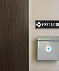 Navy Blue and White First Aid Kit sign mounted on wall above grey first aid box, displaying clear symbols for quick safety access.