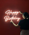 Person adjusting Neon LED Birthday Sign USB Powered Acrylic Wall Light on red wall, glowing 'Happy Birthday' text, 16.3x10.2 inches