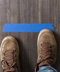 Blue painter's tape strip on dark wood floor, viewed from above with brown boots.