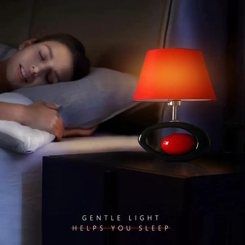 Red Modern Table Lamp With Ceramic Body And Linen Shade on a bedside nightstand next to a sleeping person, gentle light glowing.