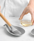 Light grey spoon rest with brush on kitchen counter, part of Three Piece Silicone Spoon Rest Set For Stove Top And Kitchen Counter