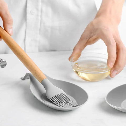 Light grey spoon rest with brush on kitchen counter, part of Three Piece Silicone Spoon Rest Set For Stove Top And Kitchen Counter