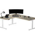 Heavy Duty L Shaped Electric Standing Desk With Smooth Glide Motors Rock Solid White Frame Spacious Bright Beech Top with laptop and monitor.