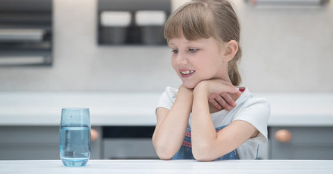 Young girl smiling at a glass of water on a table in a modern kitchen.