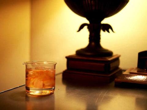 Lab Beaker Rocks Glasses Set Of 2 For Cocktails And Science Themed Barware With A Dope Scientific Twist on a table with books and lamp.