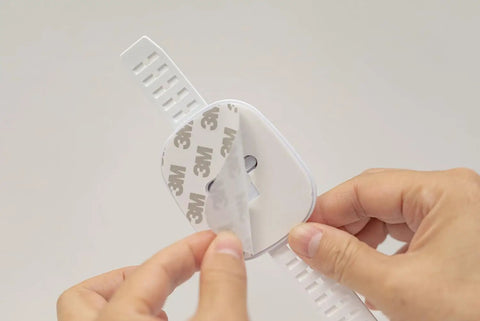 Close-up of a hand peeling adhesive on an Upgraded No Drill Furniture Wall Anchor For Baby Proofing And Child Safety, white strap lock.