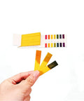 Hand holding test strips from Two Pack PH Test Strips 1 To 14 And 6.4 To 8.0 with color chart for testing water and soil.