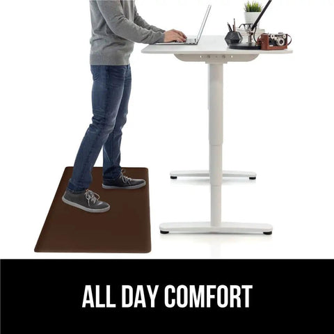 Person standing at a desk on a brown 2 Pack Cushioned Kitchen Floor Mats For Comfortable Standing 17x24 And 17x48 Waterproof And Stain Resistant One Half Inch Thick Soft Ergonomic Anti Fatigue Mat With Foam Support For Feet Brown.