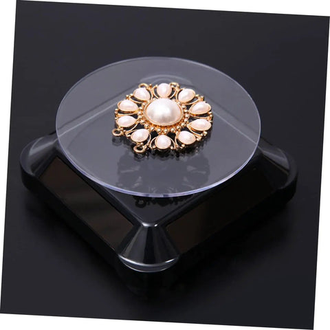 Solar Powered Rotating Jewelry Display Stand With Built In Kickstand showcasing a gold floral brooch on a clear turntable.