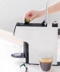 Hand inserting capsule into Compact Smart Automatic Capsule Coffee Machine With Grind And Brew Tech One Touch Espresso Maker For Your Home Office Brews Ground Or Whole Bean Coffee Keeps Coffee Hot For 40 Minutes Multi Flavor Options, with cups filled.