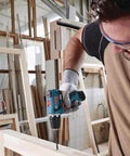 Bosch cordless drill side view in a woodshop; blue and black drill with bit, worker wearing safety glasses.