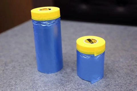 Two rolls of All Purpose Automotive Masking Tape And Plastic Sheeting Kit, blue film with yellow tape, on a gray surface.