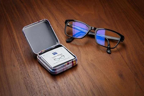 ZEISS lens cleaning tin box with hinged lid, open to reveal lens wipes; eyeglasses on a wooden surface
