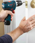 Side view of Bosch cordless drill in blue and black being used on an interior door near a brass doorknob.