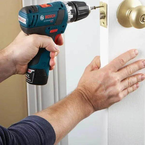 Side view of Bosch cordless drill in blue and black being used on an interior door near a brass doorknob.