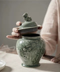 Top Tier Ceramic Airtight Tea Canisters For Loose Tea Leaves And Coffee Beans Hand Embossed Celadon Kirin Design, shown with lid being removed.