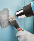Using the Powerful 1200W Heat Gun With Dual Temperature Settings to remove paint from a wall with a metal scraper.