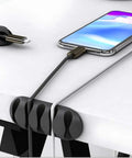 Black cable clips holding charging cables on a white desk, part of 16 Pack White Self Adhesive Cable Clips For Desk And Nightstand Cable Management.