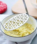 Potato masher with perforated stainless steel disk above mashed potatoes in a white bowl.
