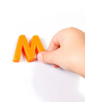 Child's hand holding orange letter 'M' magnet for Mega Size Magnetic Double Sided Kids Easel Built Tough For Endless Creative Sessions Adjustable Height Dry Erase And Chalkboard Art Station Features Four Roomy Storage Pockets Paper Roll Holder Totally Stress Free Setup Today.