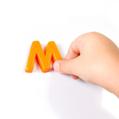 Child's hand holding orange letter 'M' magnet for Mega Size Magnetic Double Sided Kids Easel Built Tough For Endless Creative Sessions Adjustable Height Dry Erase And Chalkboard Art Station Features Four Roomy Storage Pockets Paper Roll Holder Totally Stress Free Setup Today.