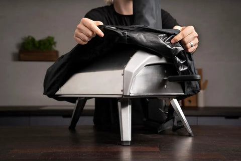 Person placing Karu 12 And Karu 2 Carry Cover Waterproof Weatherproof Shield on pizza oven, showing elastic lining and lightweight material.